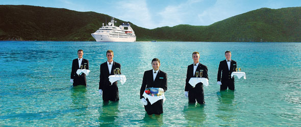 Waiters standing in the water in front of a Seabourn ship witht he line announcing it is launching the Collection a series of exclusive shore excursions