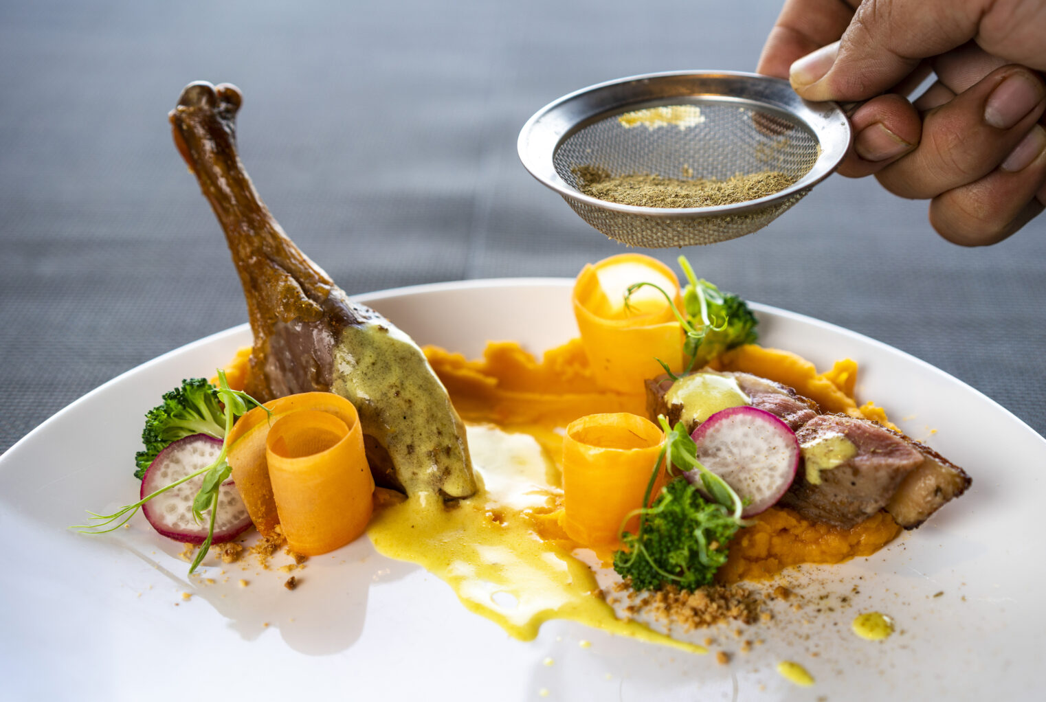 A duck leg on a plate with carrots and a person sprinkling herbs on the dish