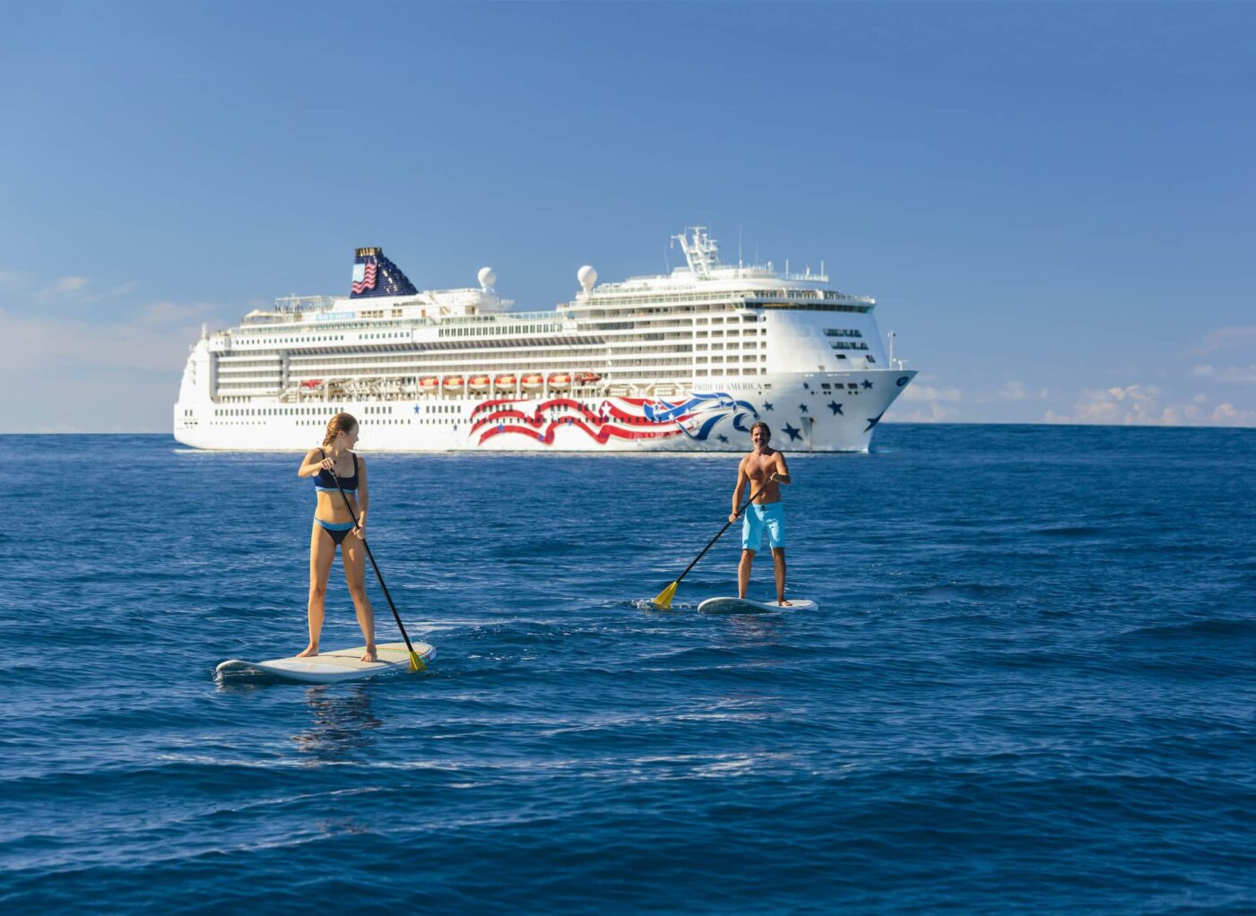 Two standup paddleboarders with an NCL ship in the background.