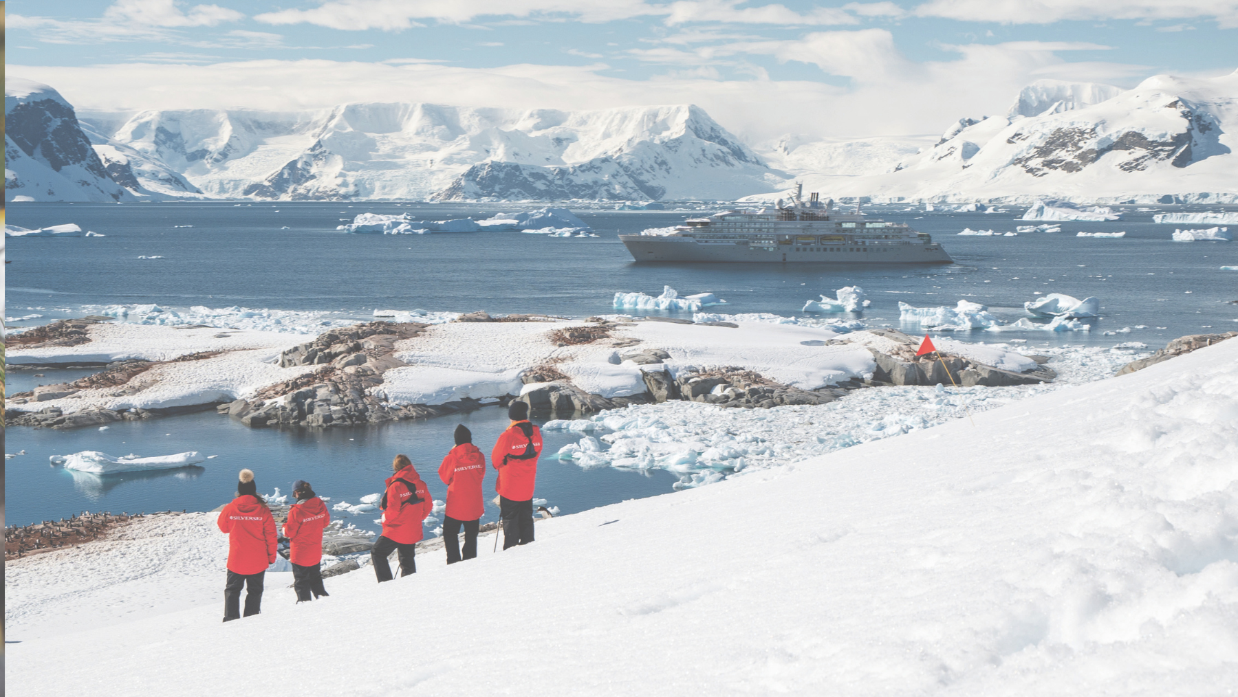 A group of people in red jackets standing on a snowy hill next to a boat