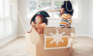 Playing, box boat and pirate children role play, fantasy imagine or pretend in cardboard container. Telescope, fun home game or sailing black kids on Halloween cruise adventure with yacht captain