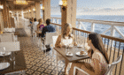 Two women dine on a ship's deck with the ocean in the background