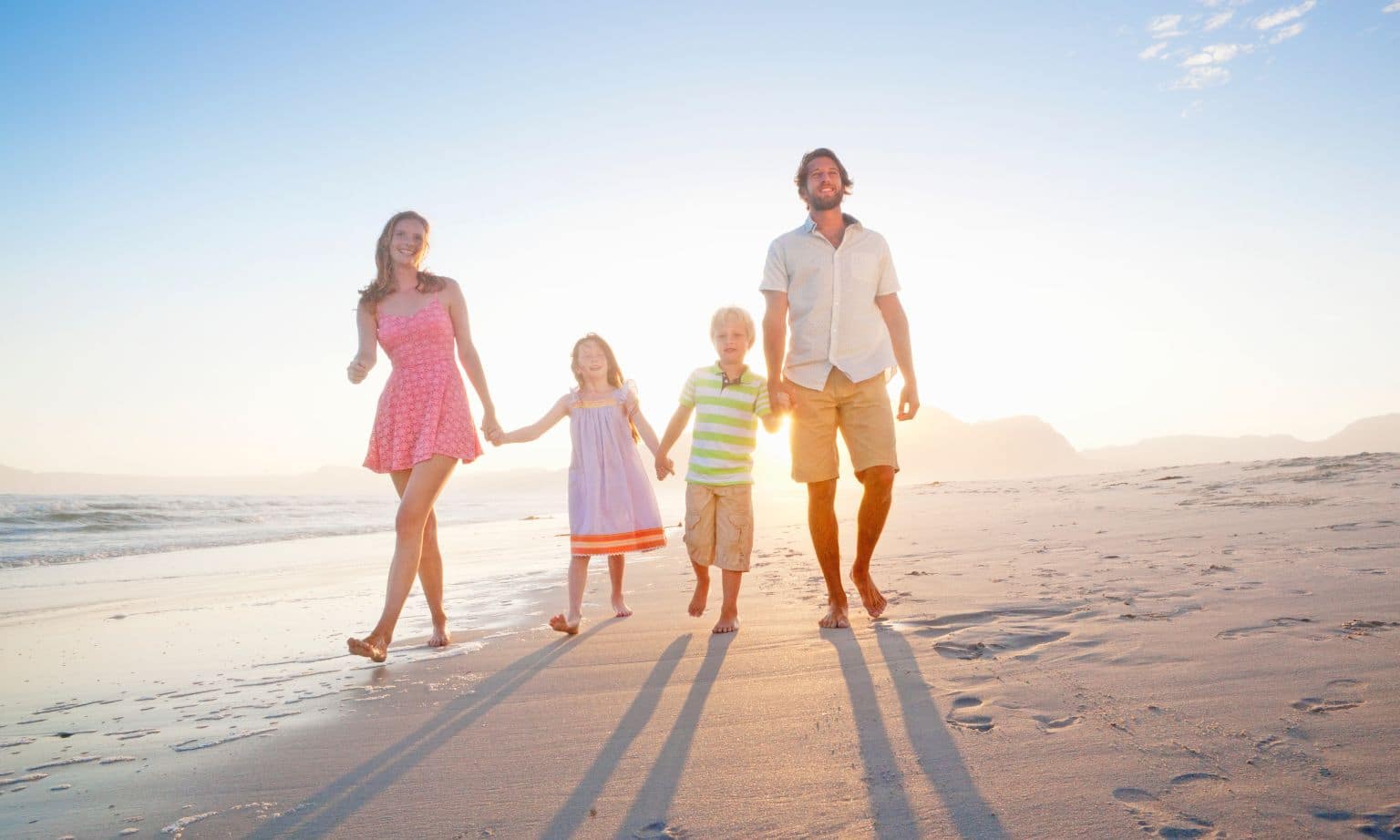 A family of two adults and two children walk along a beach holding hands