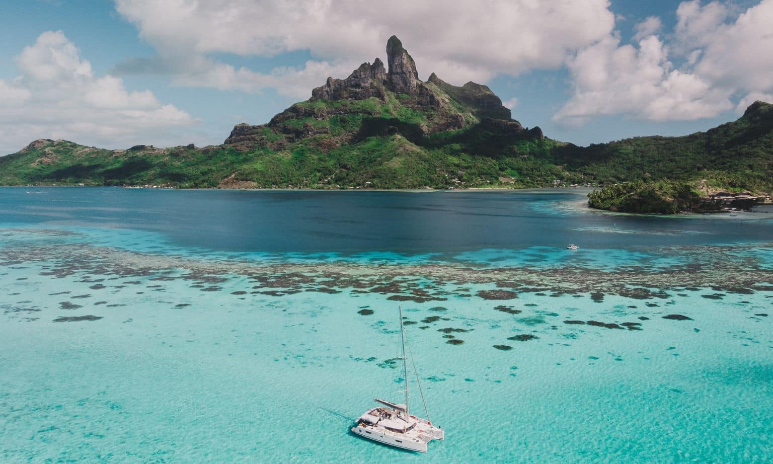 A large catamaran floats on blue water in front of a rocky island in the south pacific