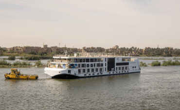 A large river cruise ship is pulled by a yellow tug down a brown river