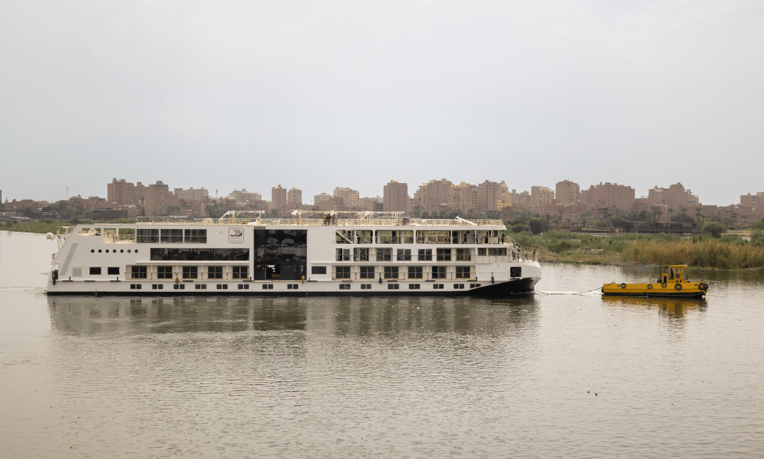 A white three storey river cruise boat is pulled down a river by a smaller boat