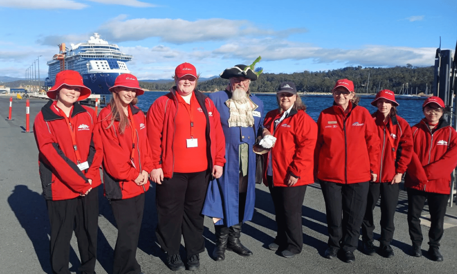 Seven people in red ambassador jackets with a town crier in the middle stand on a dock with a large cruise ship in the background.